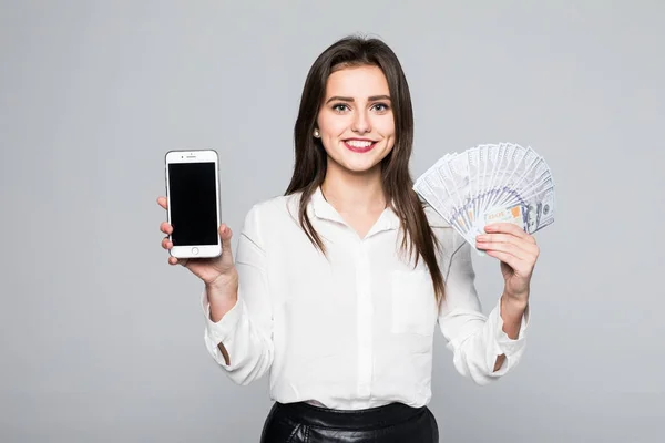 Alegre joven mujer de pie aislado sobre fondo blanco mirando cámara sosteniendo el dinero que muestra la pantalla del teléfono . —  Fotos de Stock