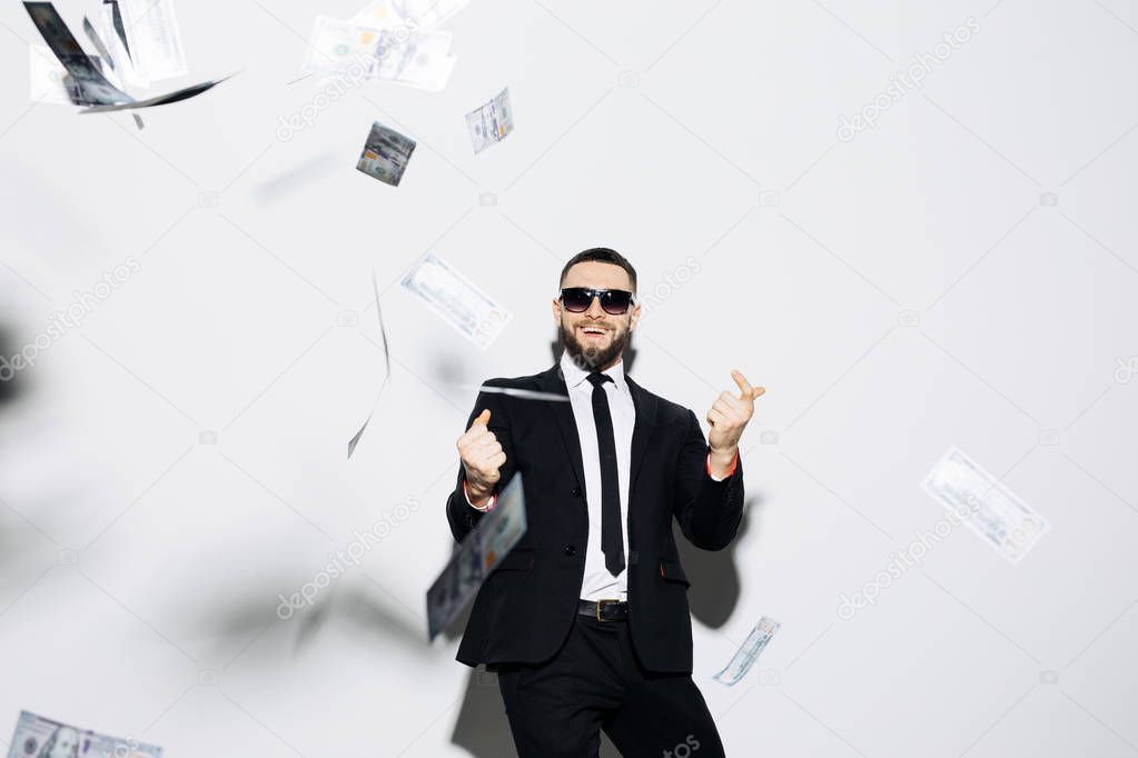 Handsome man in suit and sunglasses pointed up on fluy cash money on white background. Businessman standing in the rain of money.