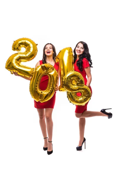 Pełnej długości portret dwóch uśmiechający się atrakcyjne dziewczyny w czerwone sukienki pozowanie, podczas gdy stojący z złoto kolorowe balony numer 2018 i patrząc na kamery na białym tle nad białym tle — Zdjęcie stockowe