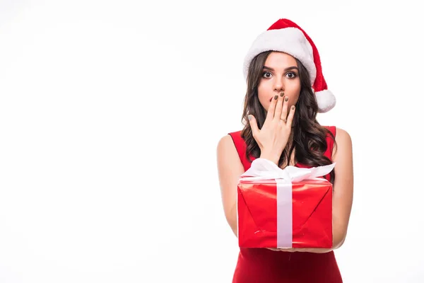 Geschokt jonge vrouw in rode jurk dragen kerst hoed staande geïsoleerd over witte muur achtergrond. Uitziende camera cadeau doos verrassing houden. — Stockfoto