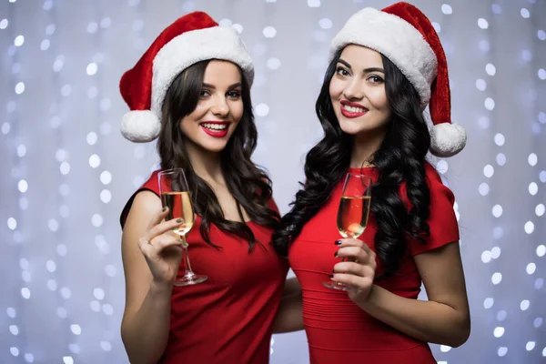 Retrato de dos mujeres en sombreros de santa y vestido rojo con champán en las manos sobre fondo claro bokeh. Vacaciones de invierno Navidad y Año Nuevo concepto —  Fotos de Stock