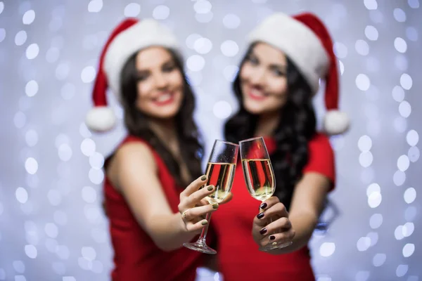 Retrato de dos mujeres con sombreros de santa y vestido rojo beben champán sobre fondo claro bokeh. Vacaciones de invierno Navidad y Año Nuevo concepto —  Fotos de Stock