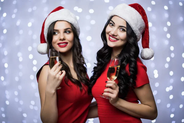 Retrato de dos mujeres en sombreros de santa y vestido rojo con champán en las manos sobre fondo claro bokeh. Vacaciones de invierno Navidad y Año Nuevo concepto —  Fotos de Stock