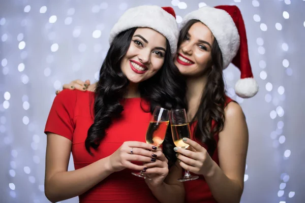 Retrato de dos mujeres con sombreros de santa y vestido rojo vítores y beber champán sobre fondo claro bokeh. Vacaciones de invierno Navidad y Año Nuevo concepto —  Fotos de Stock