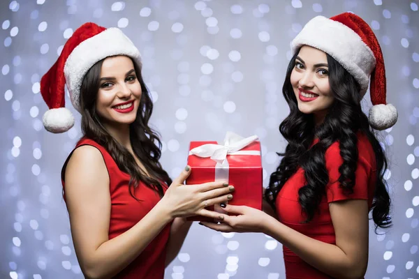 手で贈り物をサンタの帽子と赤いドレスで 2 人の女性の肖像画はボケ味の明るい背景にカメラに見えます。冬の休日のクリスマスと新年のコンセプト — ストック写真