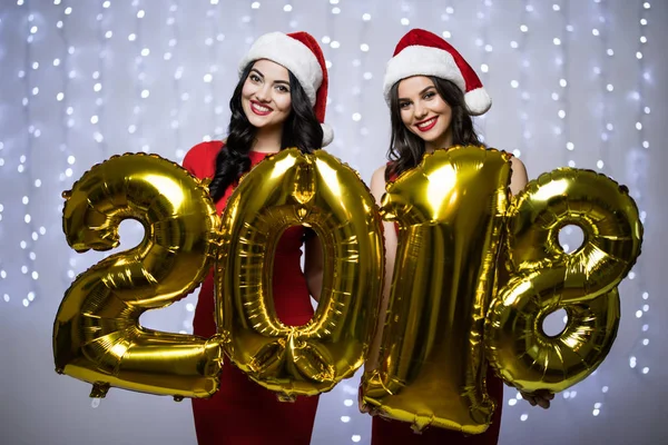 Portret dwóch kobiet w santa hat i czerwona sukienka z numerami 2018 w ręce og nowy rok na jasnym tle bokeh. — Zdjęcie stockowe