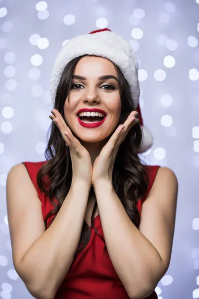 Ritratto di donna felice eccitato in cappello di Babbo Natale e rosso abito mani sul viso bokeh sfondo chiaro. Vacanza invernale Natale e Capodanno concetto — Foto Stock