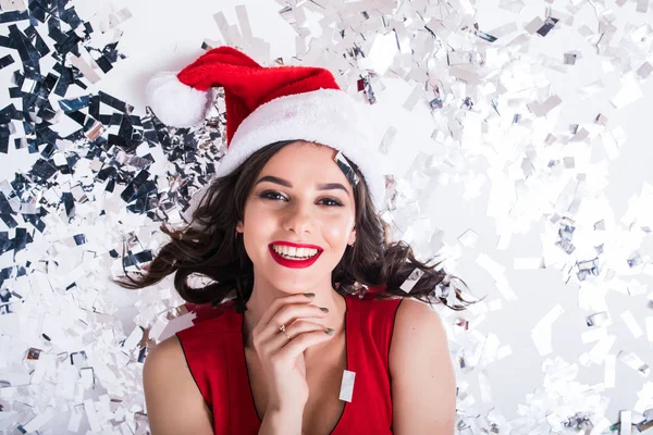 Vista superior de la mujer en rojo navidad santa claus sombrero de fiesta acostado en el suelo con confeti brillante usando vestido de noche brillante en blanco —  Fotos de Stock