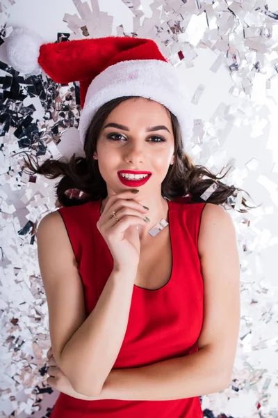 Vista dall'alto sulla donna in rosso Natale Babbo Natale cappello da vacanza sdraiato sul pavimento con coriandoli brillanti indossando abito da sera scintillante su bianco — Foto Stock