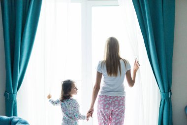 Genç anne sabah rahat yatak odasında pencere perdeleri açmayı kızı ile. Arkadan görünüş