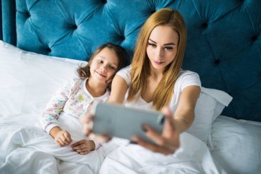 Anne ve kızı yatakta yatarken ve smartphone ile kendi kendine portre çekici. Yatak Odası, küçük bir kız ile selfie alarak kadın.