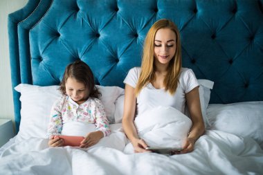 Gülümseyen anne ve kızı yatakta aygıtlarla rahatlatıcı