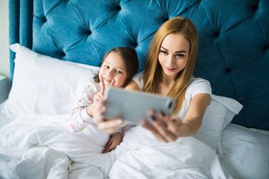 Anne ve kızı yatakta yatarken ve smartphone ile kendi kendine portre çekici. Yatak Odası, küçük bir kız ile selfie alarak kadın.