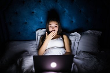 Genç güzel internet bağımlısı kadın yatakta bıkkın ve yorgun dizüstü bilgisayar ile karanlık bir odada hafif esneme yorgun düşen uyku gece geç saatlerde çalışan ev yatak odası, pijama