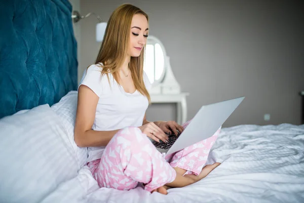 Улыбающаяся женщина наверстывает упущенное в соцсетях, расслабляясь в постели с ноутбуком в ленивый день — стоковое фото