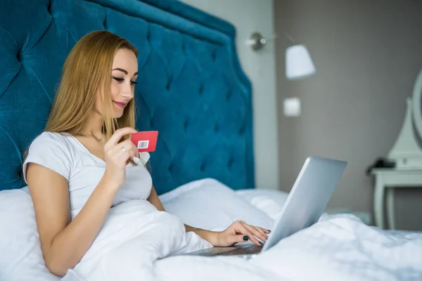 Улыбающаяся женщина лежит на кровати с кредиткой в руке, когда она использует свой ноутбук, когда просыпается утром — стоковое фото