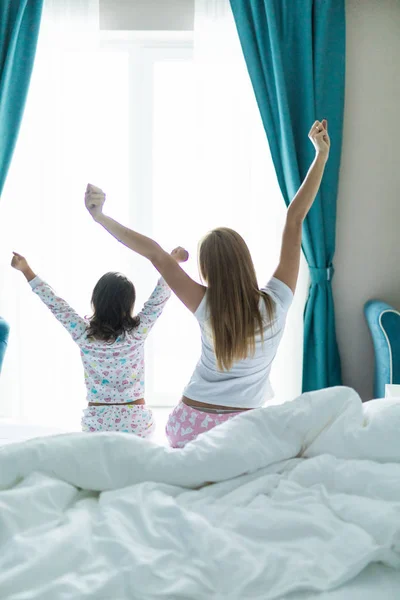 Обратный вид матери и дочери растягивается и зевает, сидя на кровати дома — стоковое фото