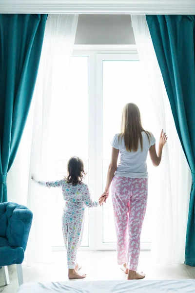 Молодая мама с дочерью открывают окна шторы в уютной спальне по утрам. Вид сзади — стоковое фото