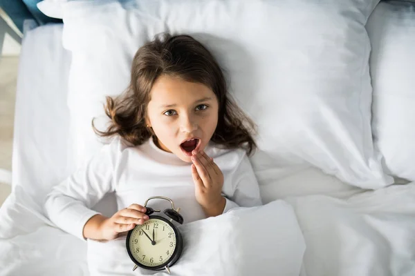 Top view of amazed girl covering her mouth with hand, lying in bed with alarm clock at home, looking at camera — Stock Photo, Image