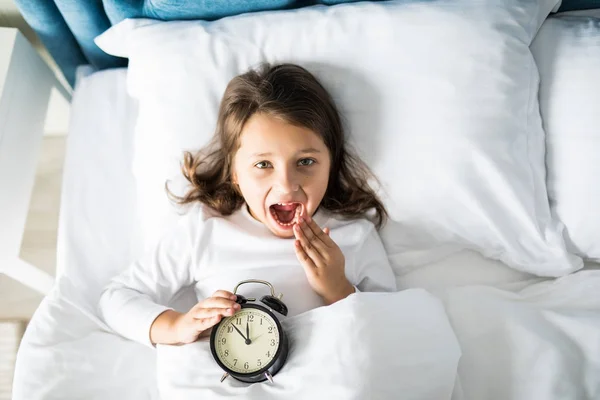 Top view of cheerful girl lying in bed with alarm clock at home, looking at camera — Stock Photo, Image