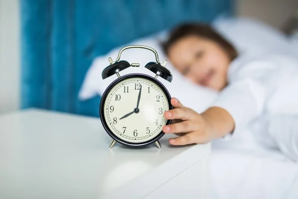 Portrait of a shocked cute little girl holding alarm clock and looking at camera while lying in bed — Stock Photo, Image