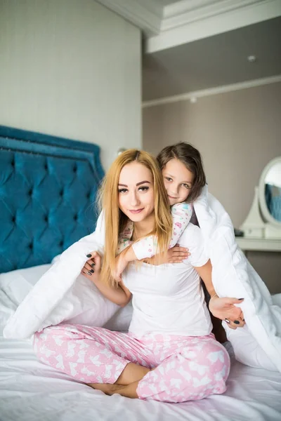 Πορτρέτο του μελαχρινή μακριά μαλλιά μητέρα και κόρη κάτω από το πάπλωμα που συμμετέχουν στο μαλακό φως στο λευκό λινό κρεβάτι το πρωί. Έννοια της ευτυχισμένη οικογενειακή ζωή, χαλάρωση, άνεση, διασκέδαση. Κάτοψη. — Φωτογραφία Αρχείου