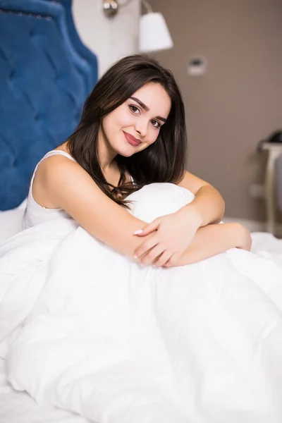 Gyönyörű fiatal nő mosolyogva ül ágyban reggel hálószoba. — Stock Fotó