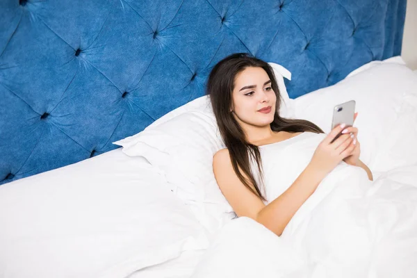 Feliz joven mujer acostada en la cama y mensajes de texto en el teléfono inteligente en el dormitorio en casa —  Fotos de Stock