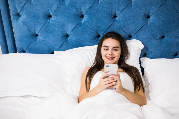 Счастливая молодая женщина, лежащая в постели и текстурирующая на смартфоне в домашней спальне — стоковое фото