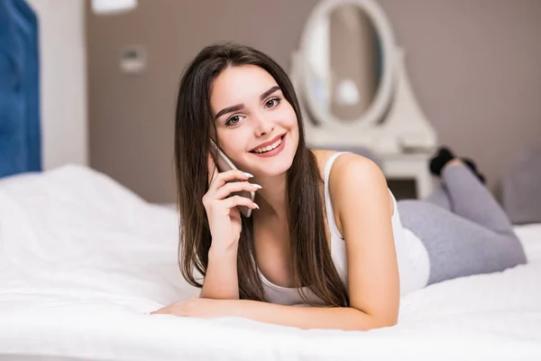 Hermosa mujer rubia hablando en su teléfono tendida en su cama en pijama, sonriendo en casa —  Fotos de Stock