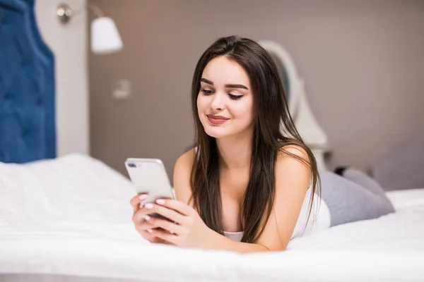 Feliz joven mujer acostada en la cama y mensajes de texto en el teléfono inteligente en el dormitorio en casa —  Fotos de Stock