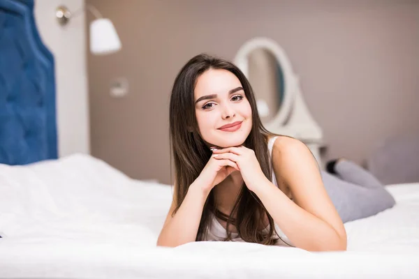 Una mujer acostada al final de la cama debajo de la colcha y sonriendo, con la cabeza apoyada en la mano con la otra en el pelo en casa —  Fotos de Stock