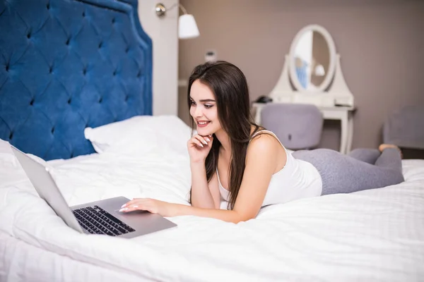 Mujer bastante sonriente ocupada navegando en el ordenador portátil mientras toma una taza de café en la cama blanca . —  Fotos de Stock