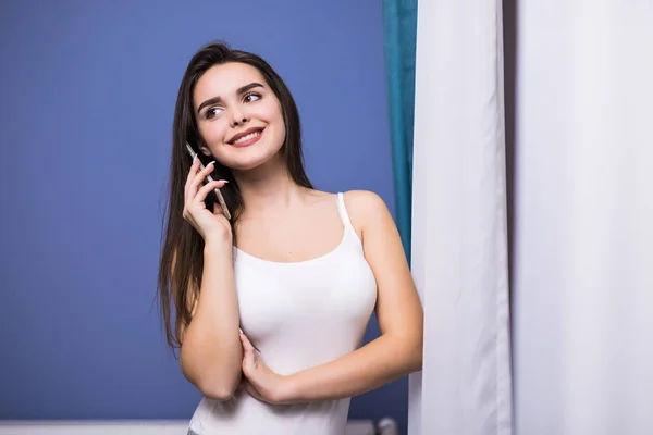 집에서 커튼에 휴대 전화로 이야기 하는 아름 다운 젊은 여자 — 스톡 사진