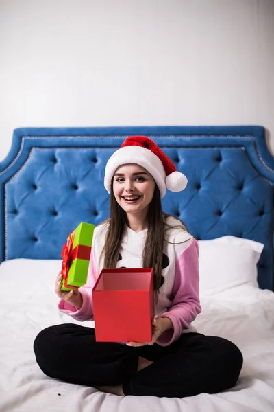 Feliz mulher sorridente em um chapéu de Natal vermelho abrir um presente na cama em casa — Fotografia de Stock