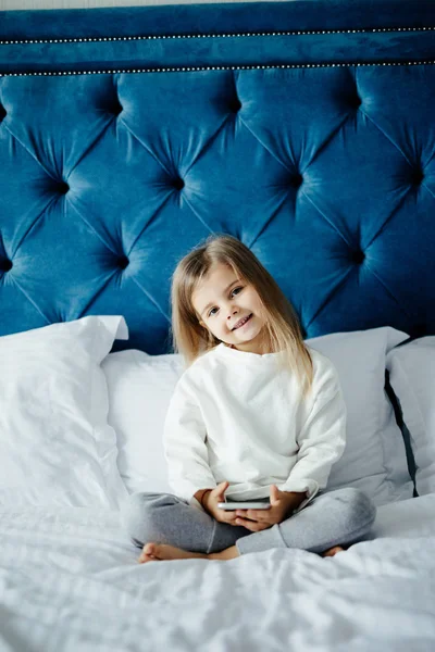Красавица маленькая девочка лежит в постели со смартфоном дома — стоковое фото
