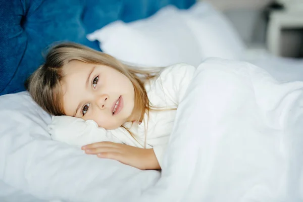 Close-up portrait of beautiful little kid lying on bed with hand under pillow, looking at camera at home — Stock Photo, Image