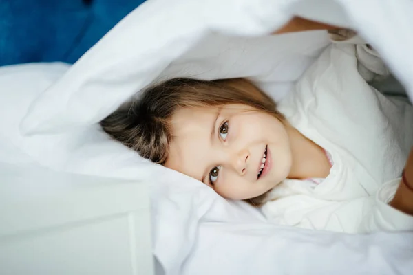 Красивая маленькая девочка в сонной одежде лежит под одеялом в постели дома — стоковое фото