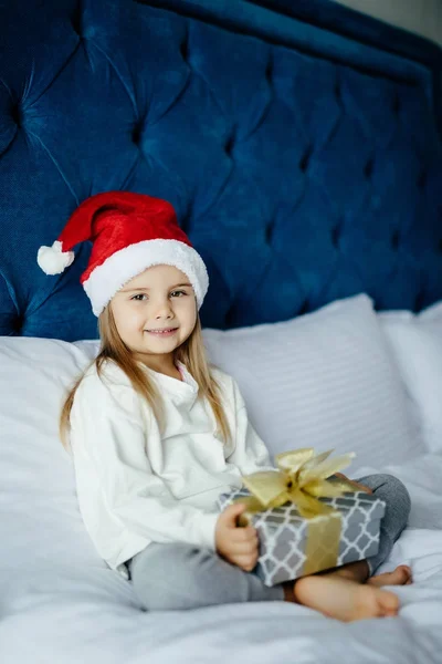 Hediye kutusu, holding Noel Baba'nın şapkalı küçük kız yatakta otururken kameraya bakarak mutlu heyecanlı — Stok fotoğraf