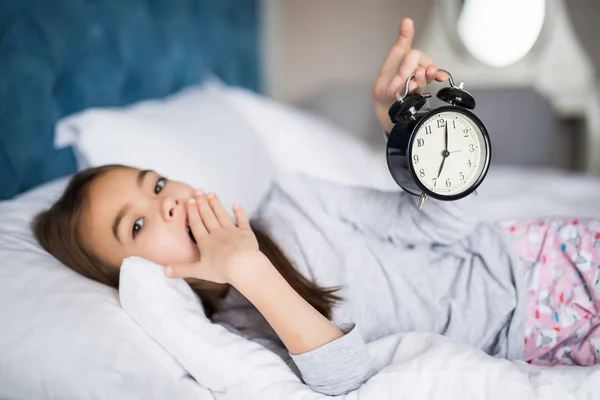 Portrait of shocked cute little girl holding alarm clock and looking at camera while lying in bed in the morning — Stock Photo, Image