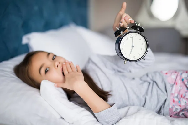 Amazed little girl holding alarm clock, covering her mouth while lying in soft bed — Stock Photo, Image