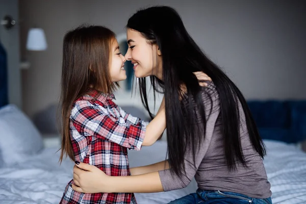 Hermosa mujer y su linda hijita están tocando las narices y sonriendo mientras están sentados en la cama — Foto de Stock