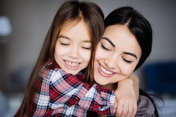 美丽的年轻母亲和她的女儿是拥抱，微笑着坐在一起在家里的床上闭着眼睛 — 图库照片