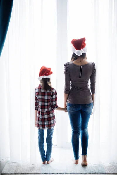 Genç anne ile kızı açılış pencere, Noel Baba şapkaları sabah Noel sabahları rahat yatak odasında Perdeleri açın. — Stok fotoğraf