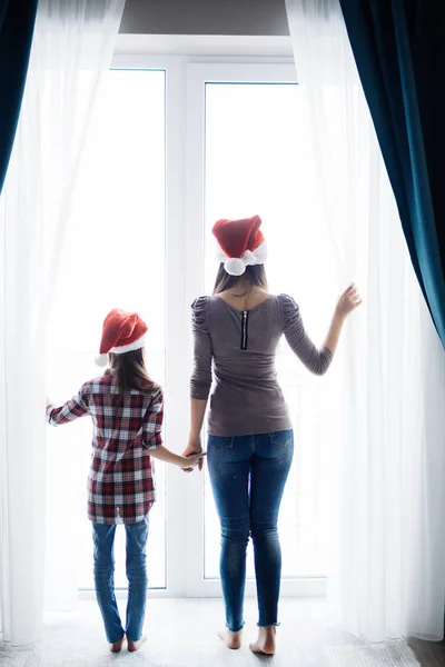 Genç anne ile kızı açılış pencere, Noel Baba şapkaları sabah Noel sabahları rahat yatak odasında Perdeleri açın. — Stok fotoğraf