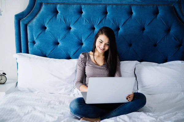 Mujer sonriente poniéndose al día en sus redes sociales mientras se relaja en la cama con una computadora portátil en el día — Foto de Stock