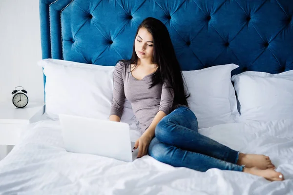 Улыбающаяся женщина наверстывает упущенное в социальных сетях, расслабляясь в постели с ноутбуком в день — стоковое фото