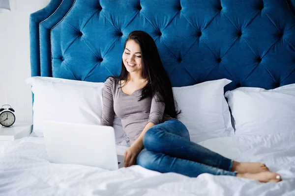 Mujer sonriente poniéndose al día en sus redes sociales mientras se relaja en la cama con una computadora portátil en el día — Foto de Stock
