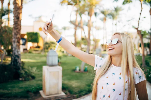 Vidám fiatal nő, kalap és napszemüveg vesz selfie mobiltelefon nyári üdülőhely pálmák háttér. — Stock Fotó