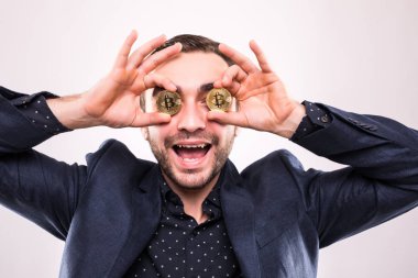 Genç yakışıklı heyecanlı adam gri arka plan üzerinde izole bitcoin paralar ile göz kapağı